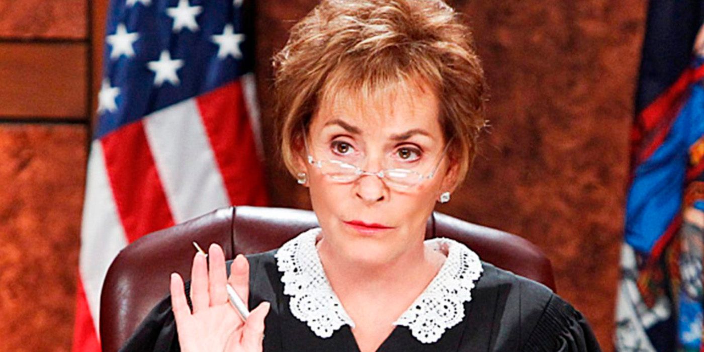 Judge Judy with her hand up, glasses on the tip of her nose with the American flag in the background.