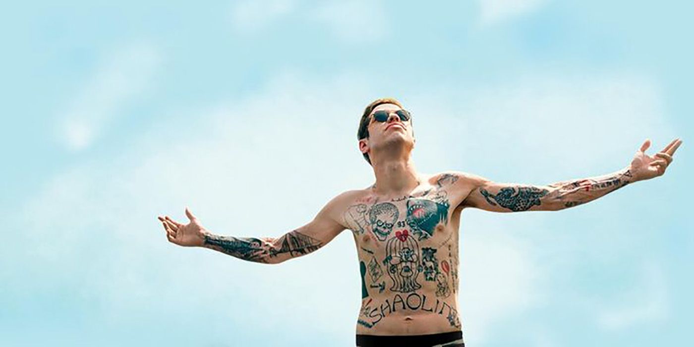 Pete Davidson wearing sunglasses, shirtless, hands in the air against a blue sky.