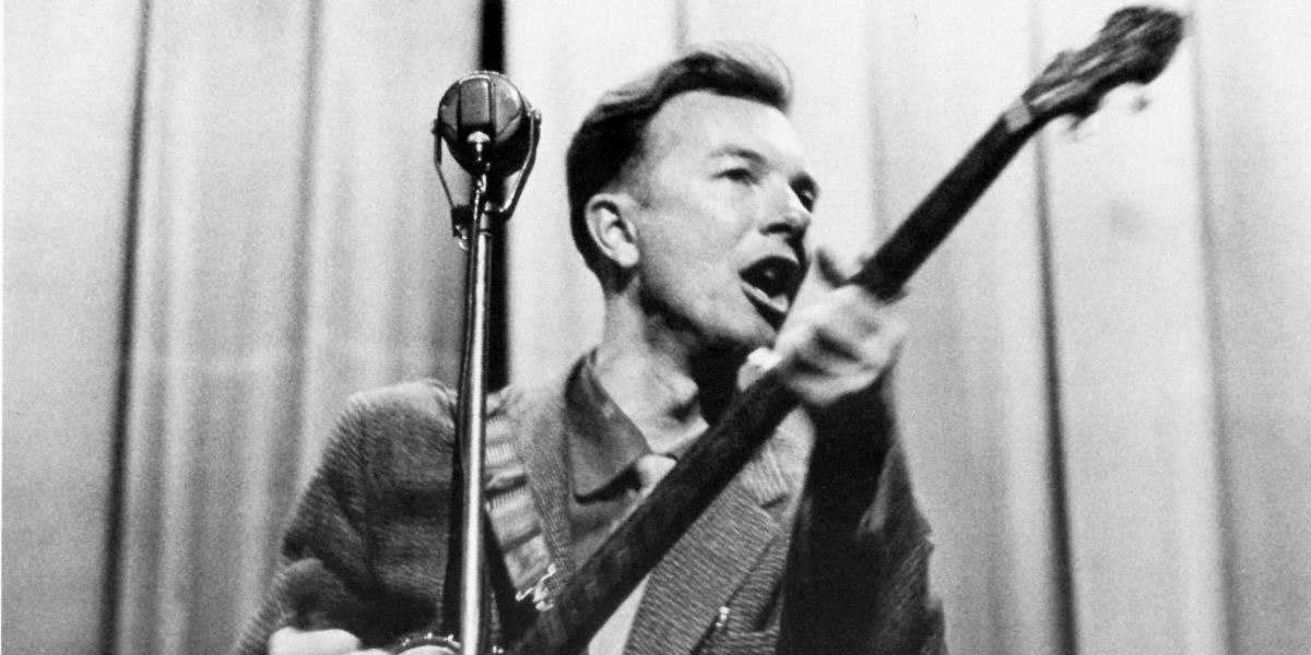 Pete Seeger bernyanyi sambil memainkan banjo di depan tirai