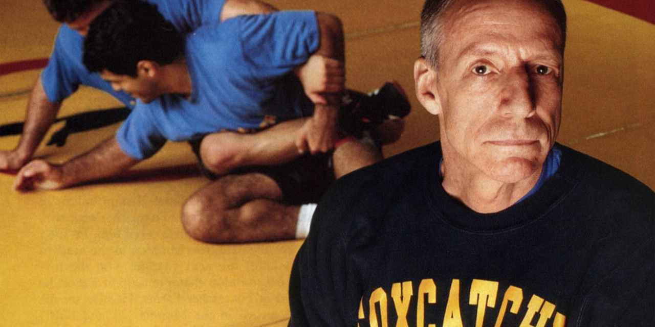 An old man stares at the camera as wrestlers fight on the floor in Team Foxcatcher