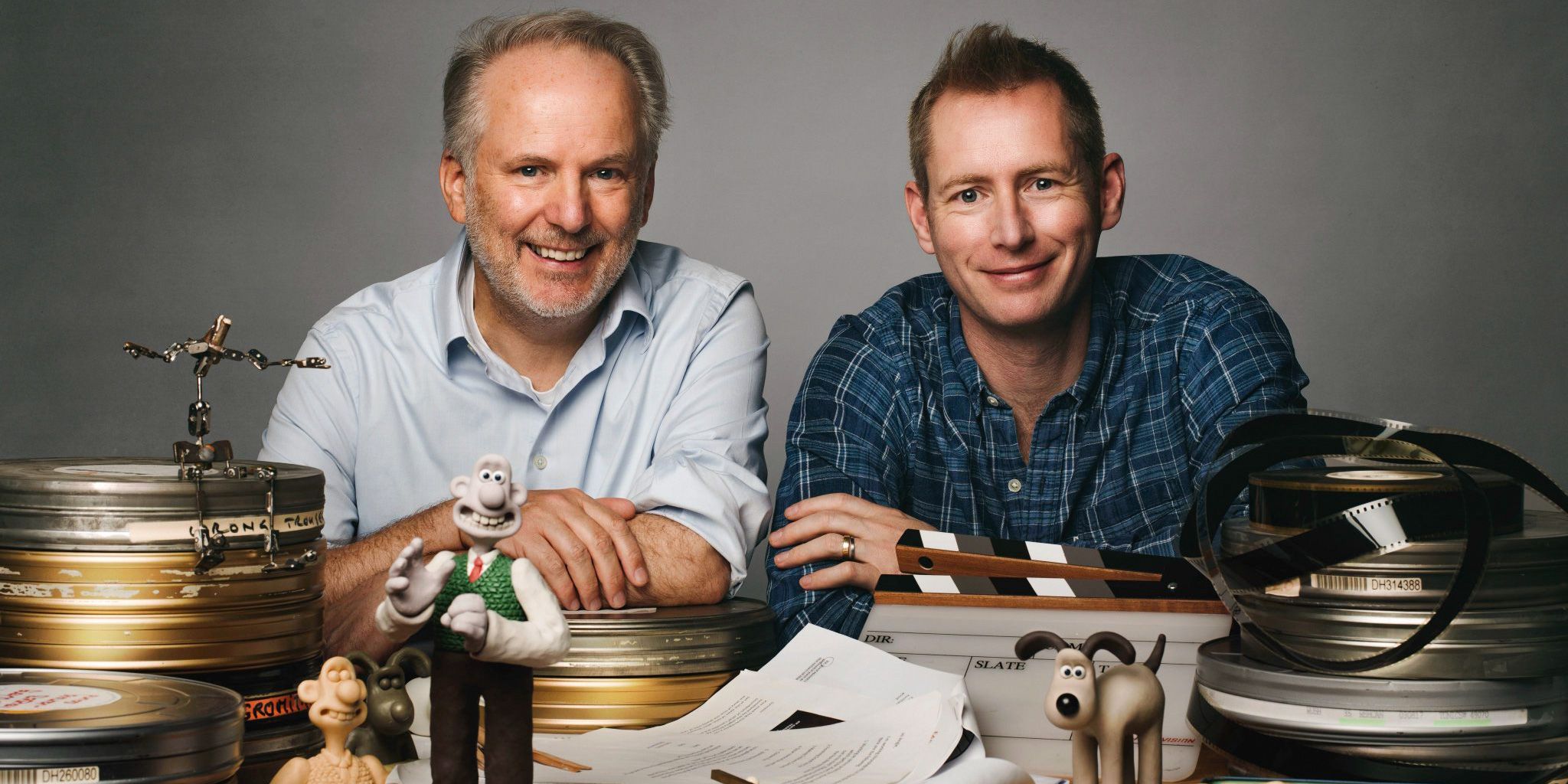 Nick Park smiles behind a table, where his creations Wallace and Gromit are stood on a table.