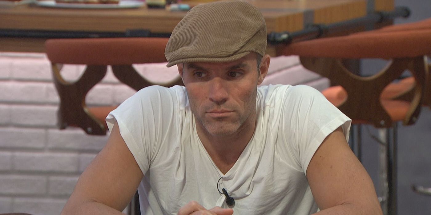 Enzo wearing a paigeboy hat and white T-shirt, sitting in the kitchen on Big Brother looking pensive.