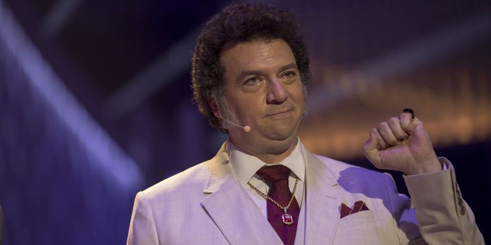 Jesse wears a suit, tie, and microphone on stage in The Righteous Gemstones