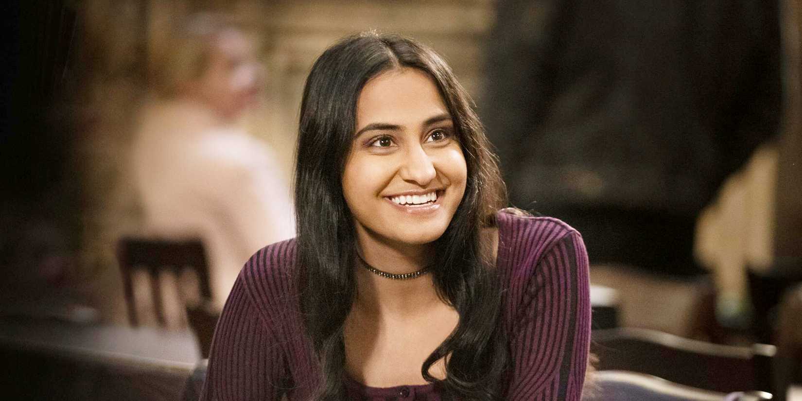Amrit Kaur as Bela Malhotra in Sex Lives of College Girls sitting and smiling into the camera in the dining hall at Essex.