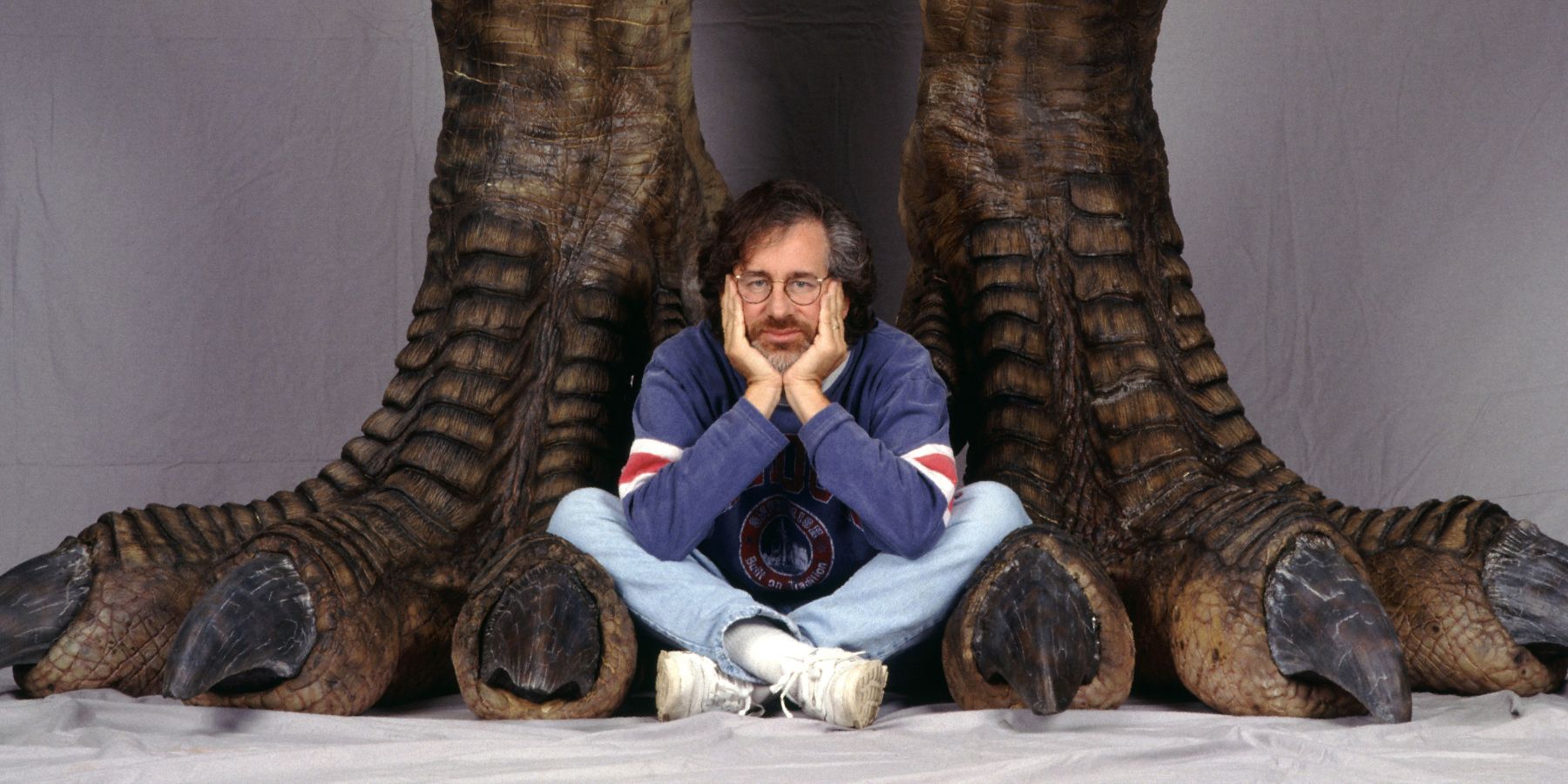 Steven Spielberg sitting between the feet of a T Rex in a promo image for Jurassic Park