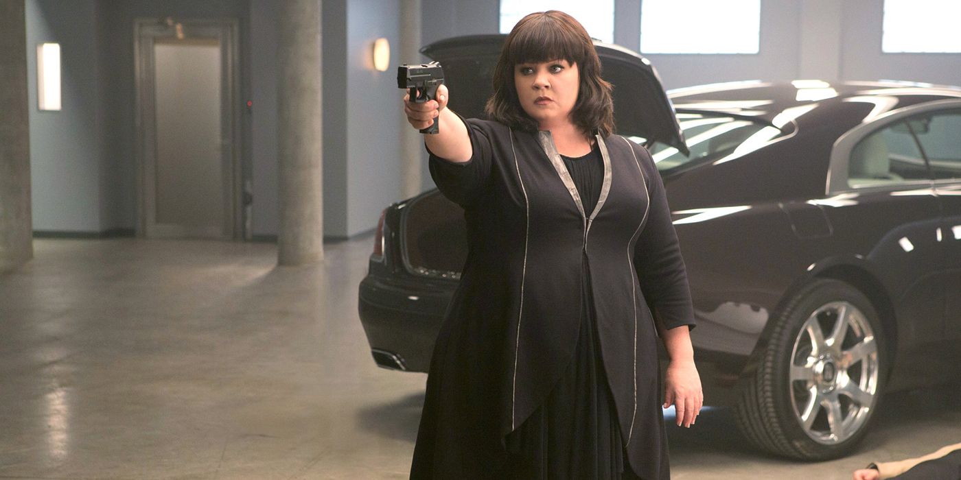 Melissa McCarthy vestida de negro, sosteniendo y apuntando con un arma a un espía.