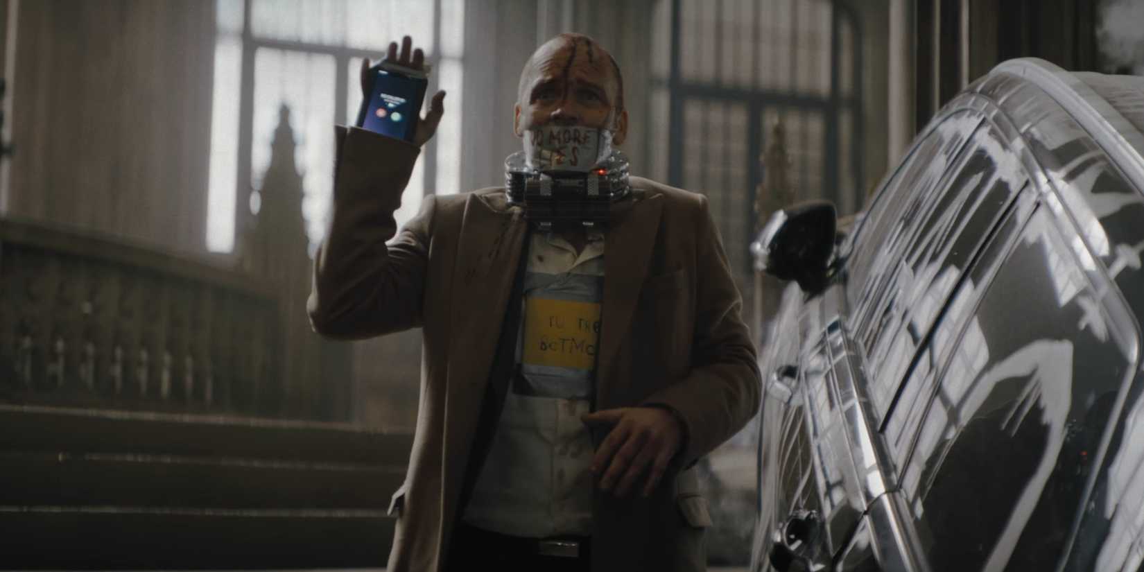 Gil Colson with a bomb around his neck holding up a phone in The Batman