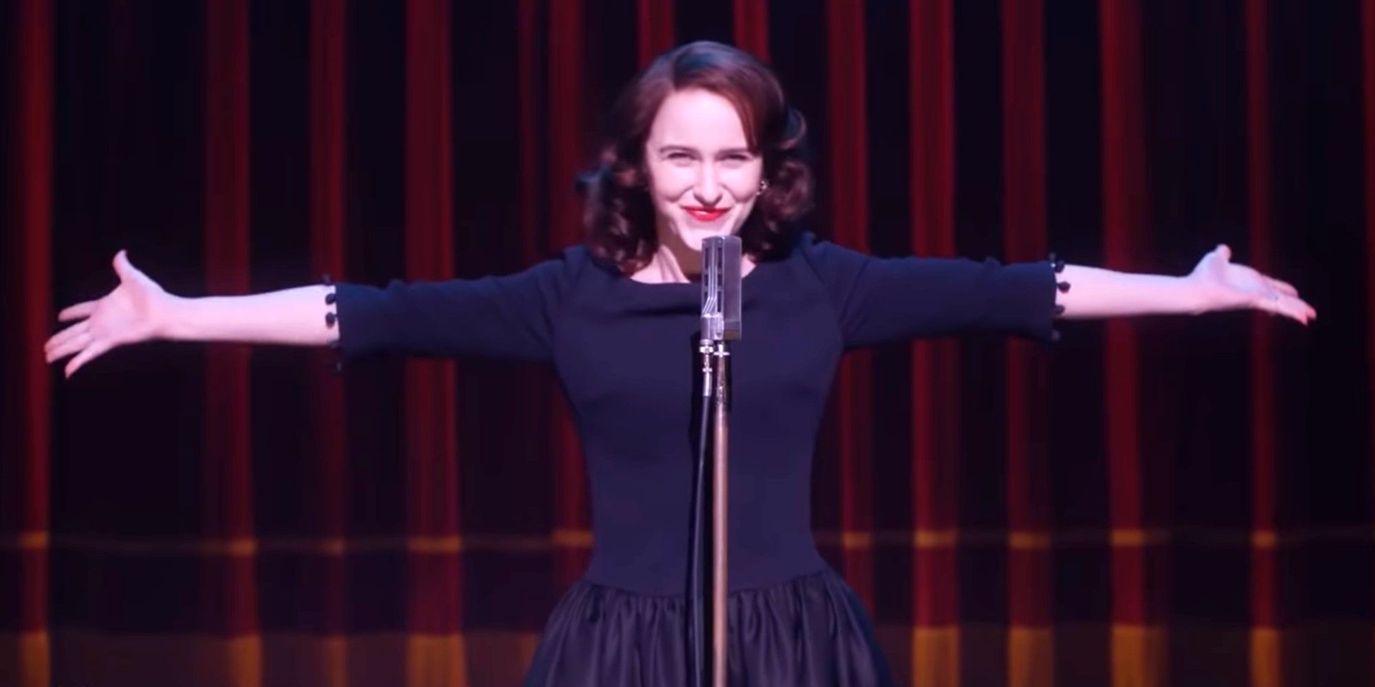 Mrs. Maisel stands in front of a microphone