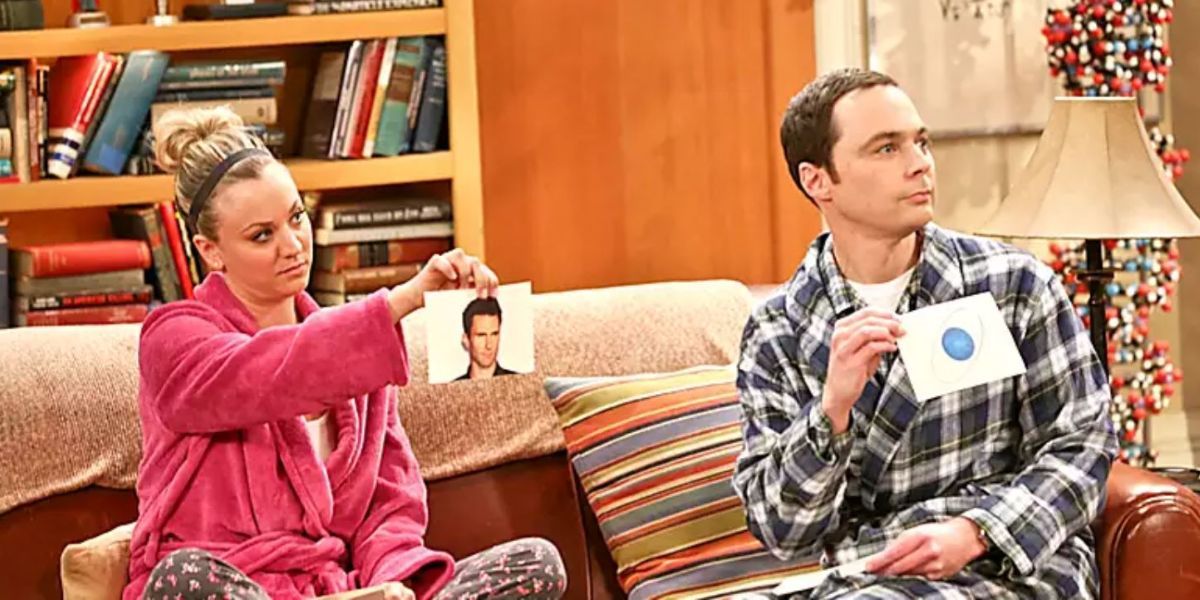 Sheldon and Penny sitting on the couch holding up cards with images in the Big Bang Theory