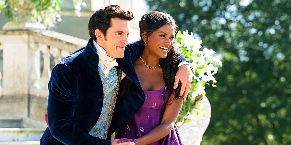 Kate (Simone Ashley) and Anthony (Jonathan Bailey) holding each other from the side while smiling in front of a flight of outdoor stairs.