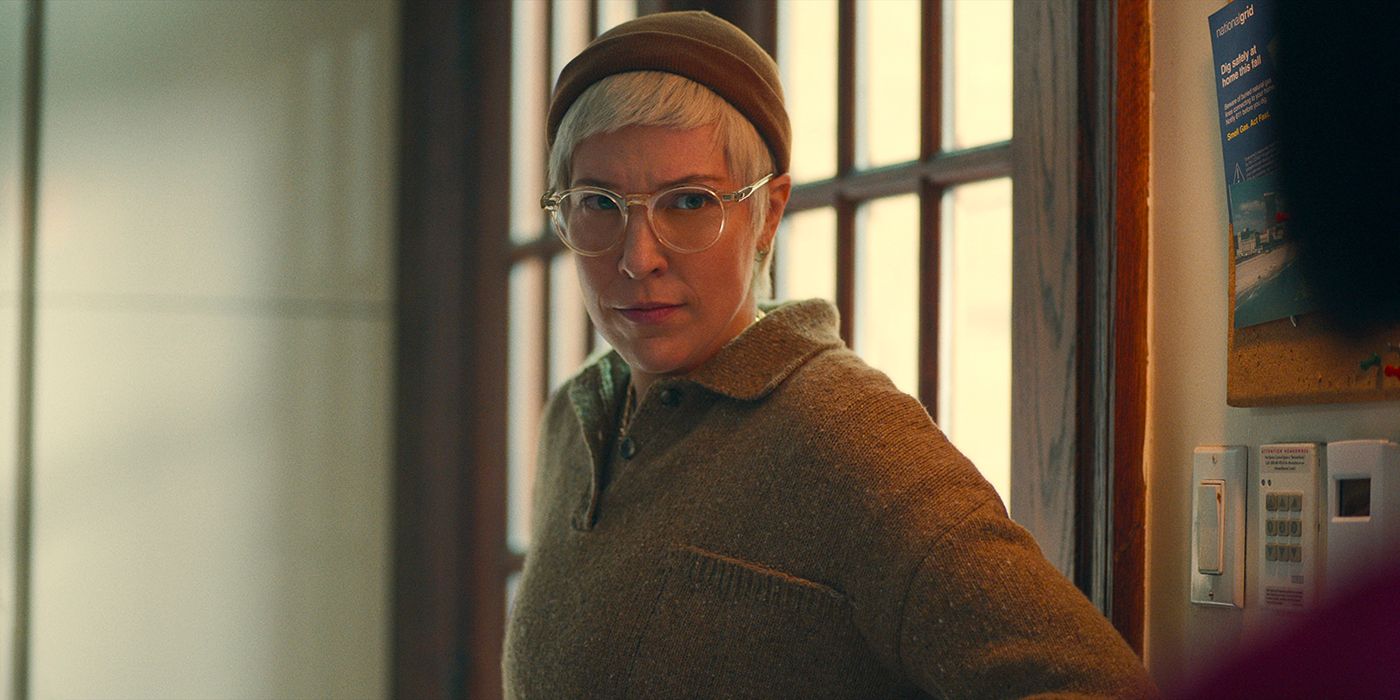 Lizzy from Russian Dolls wearing a beanie hat and glasses, standing at a doorway.
