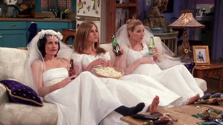 Monica Rachel and Phoebe sitting on the couch in wedding dresses and eating popcorn in Friends