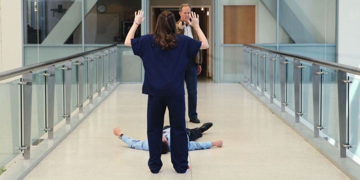 April Kepner with her back to the camera and hands up, in front of Derek Shepherd lying injured on the ground, and Gary Clark aiming a gun at April on Grey's Anatomy 