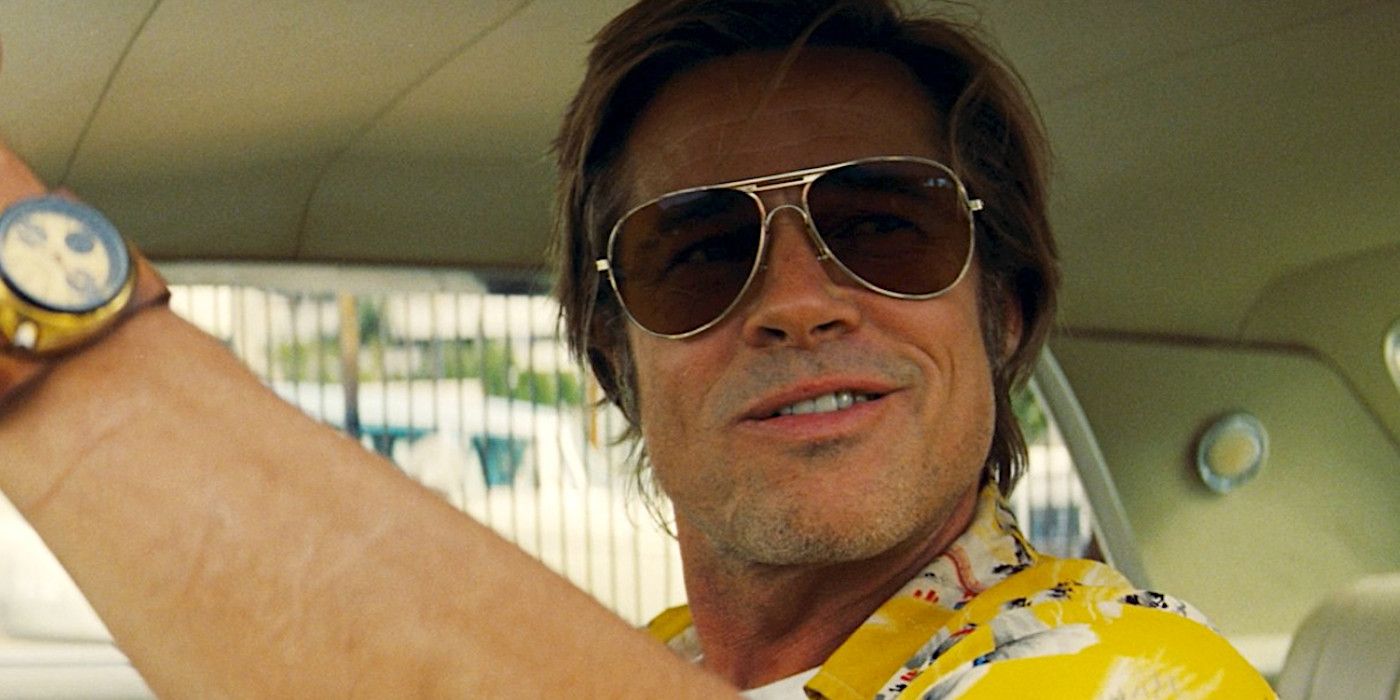 Brad Pitt in character in Once Upon a Time in Hollywood wearing a yellow shirt and sunglasses and a watch sitting behind the wheel of a car