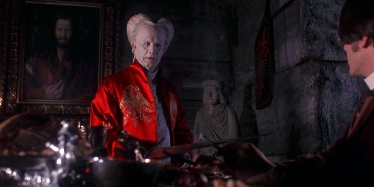 Dracula standing, wearing a red jacket with stone walls and a statue behind him.