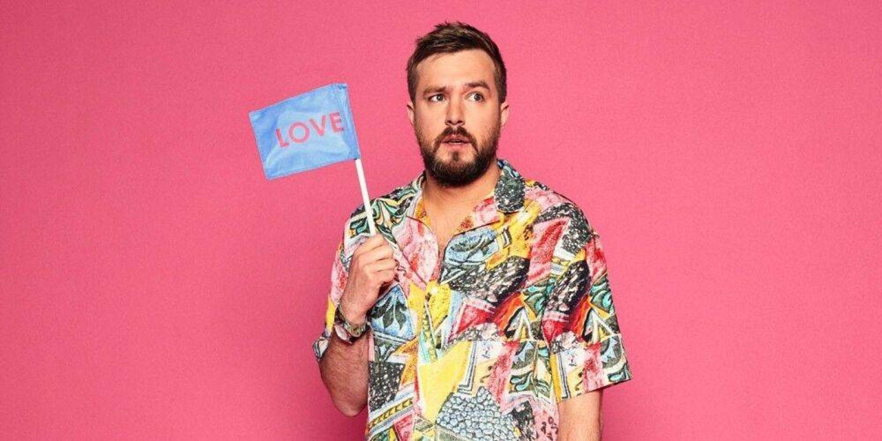 Love Island narrator Iain Sterling holds a small blue flag that reads &quot;love&quot; in pink letters.