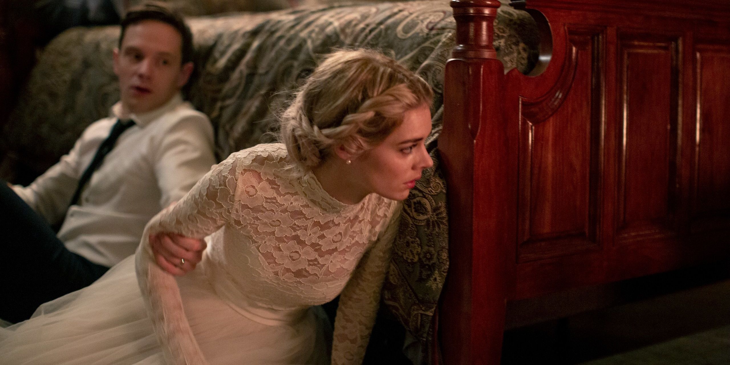 Grace hiding behind a bed with a man beside her grabbing her arm as she looks out for the killer