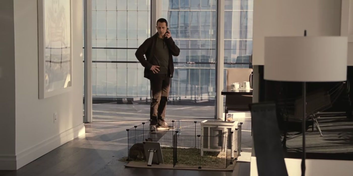 Kendall from Succession standing in his apartment on the phone looking at a bunny cage on the table.