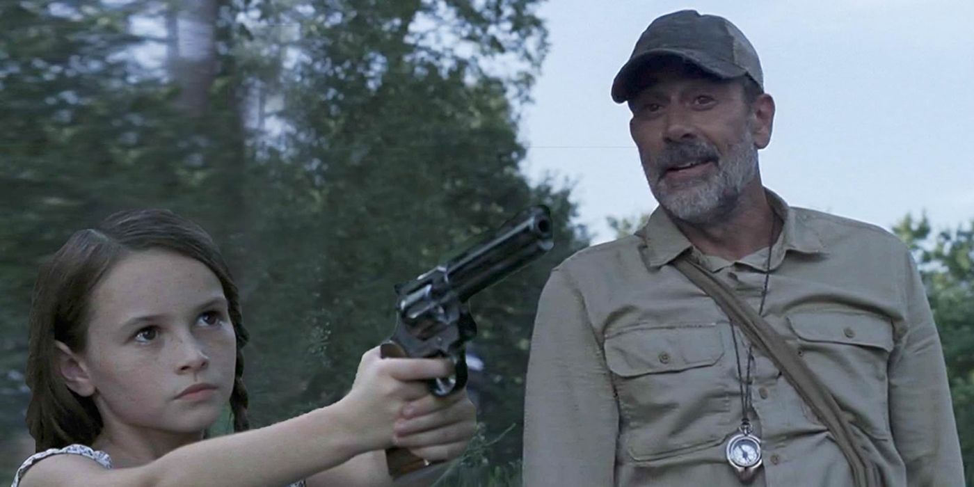 Negan from The Walking Dead wearing a baseball hat and smiling looking at Judith holding and aiming a gun.