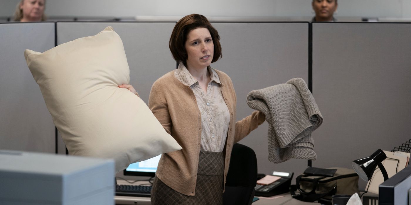 Evie Russell holding up a pillow and a blanker in her cubicle in What We Do in the Shadows.