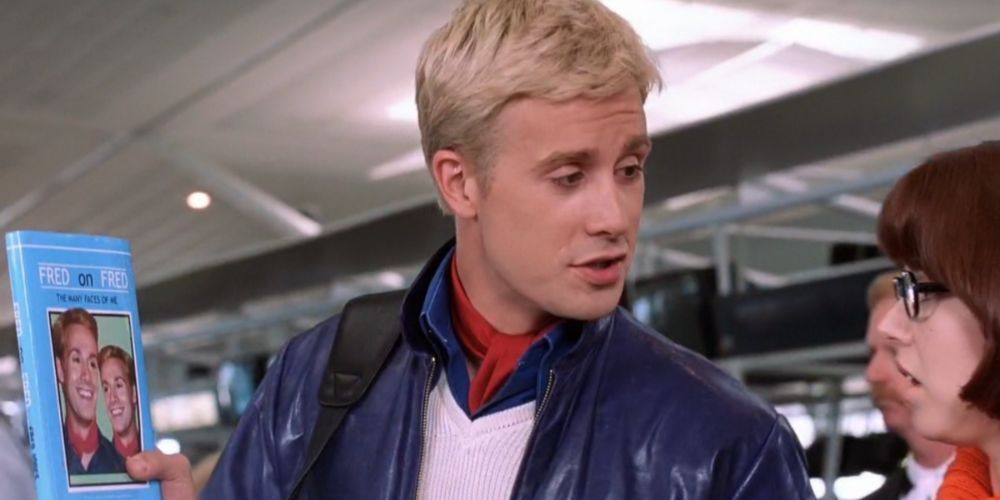 Freddie Prinze Jr as Fred stands in the airport flaunting his new book