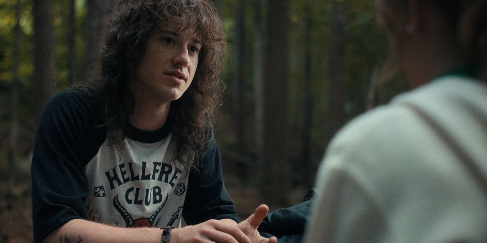 Joseph Quinn as Eddie Munson in Stranger Things season 4 with bushy, curly hair sitting in a forest