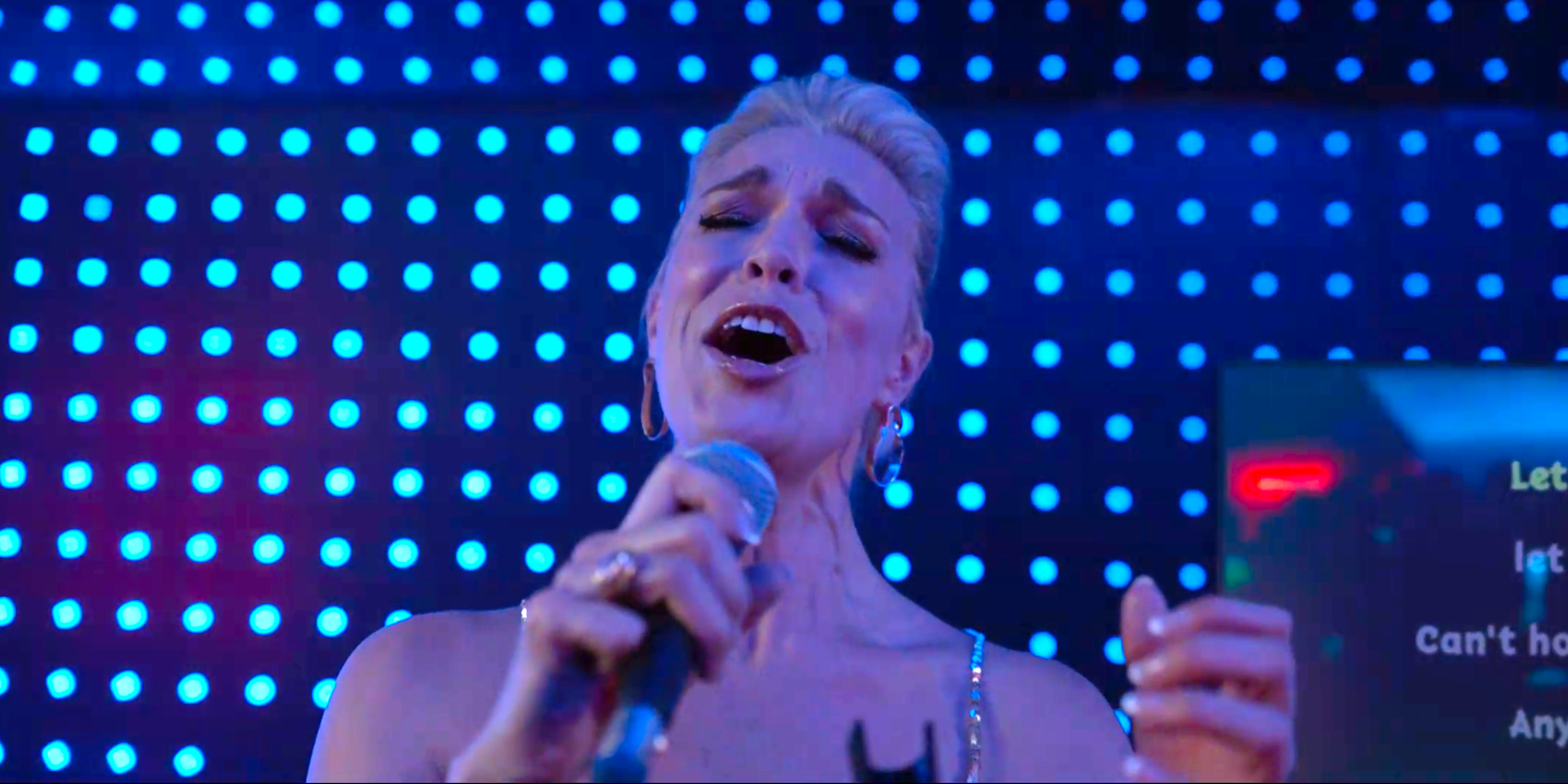 A blonde woman holds a microphone and sings in front of a blue stage
