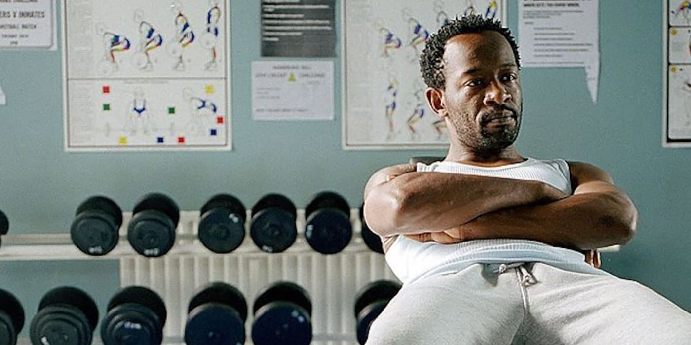 Lennie James from the British series Buried sitting, arms folded in front of rows of dumbbells.