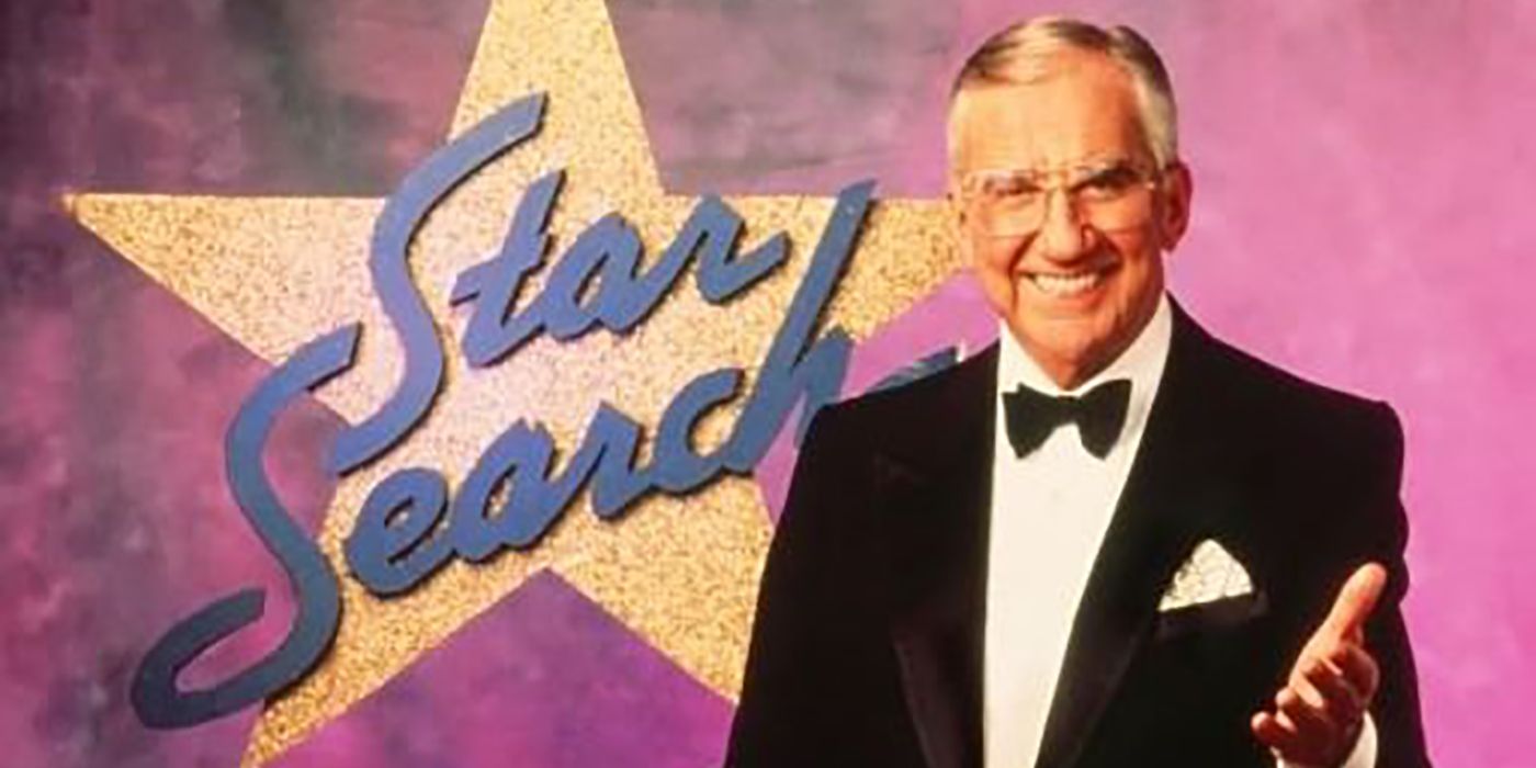 Ed McMahon standing beside the Star Search symbol, smiling with his hand up.