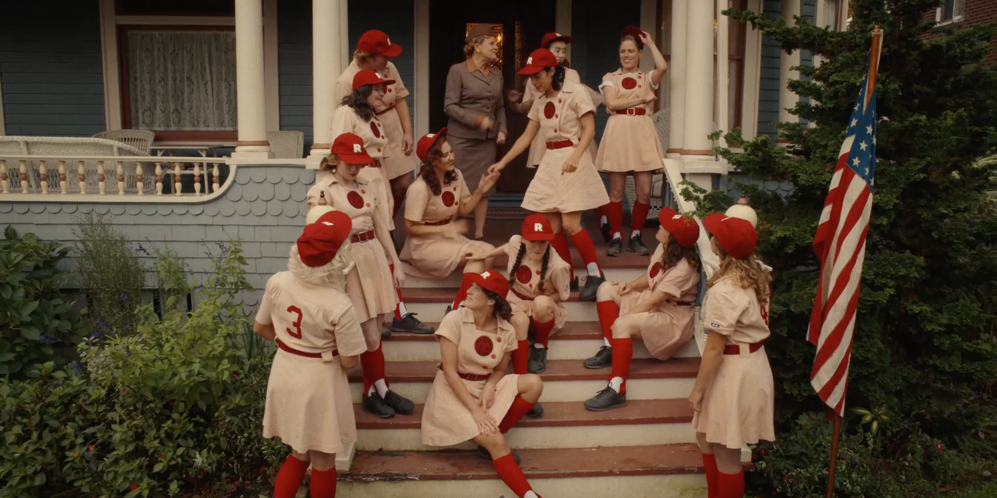 A group of women dressed in old-fashioned baseball uniforms sit on the steps of a house