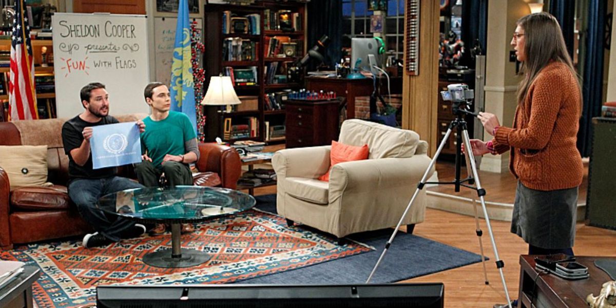Amy em pé atrás da câmera conversando com Wil e Sheldon que estão sentados no sofá segurando uma bandeira em TBBT