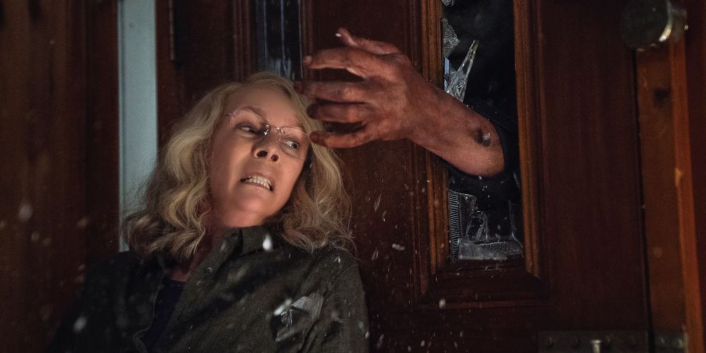 Laurie Strode contra a porta enquanto uma mão tenta abri-la por uma janela quebrada no Halloween de 2018.
