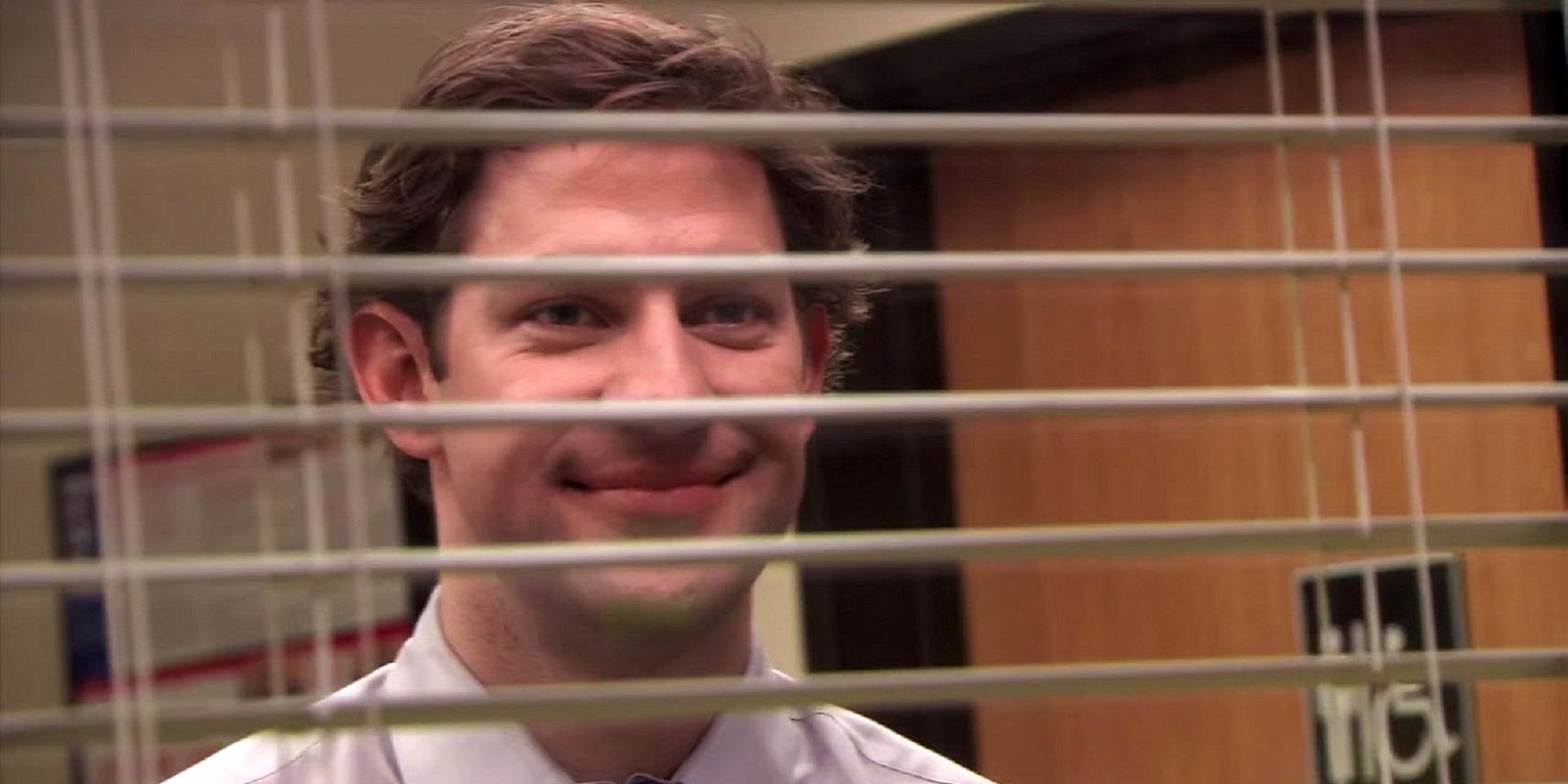 John Krasinski as Jim Halpert looking through window blinds and smiling in The Office