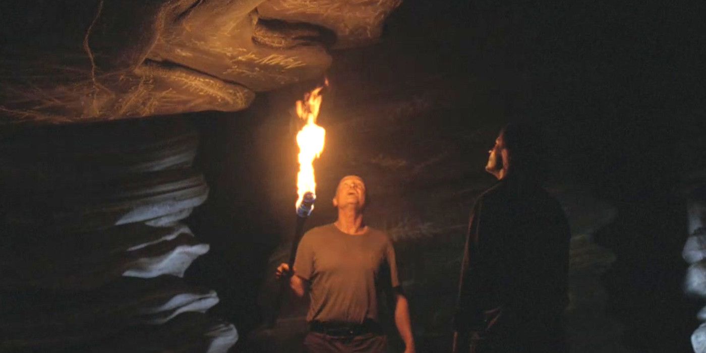 John Locke segura uma tocha e olha para uma escrita no teto de uma caverna enquanto Sawyer observa a 6ª temporada de Lost.