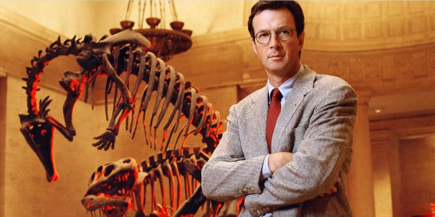 A man in a gray suit and glasses stands in front of a Tyrannosaurus rex skeleton.