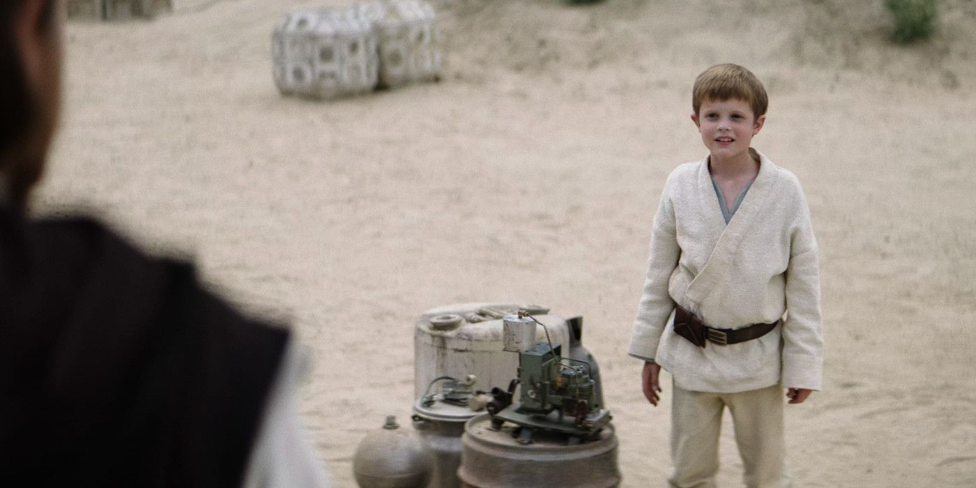 Shot over the shoulder of Obi-Wan Kenobi at young Luke Skywalker in white attire standing next to a piece of dismantled electronic equipment.