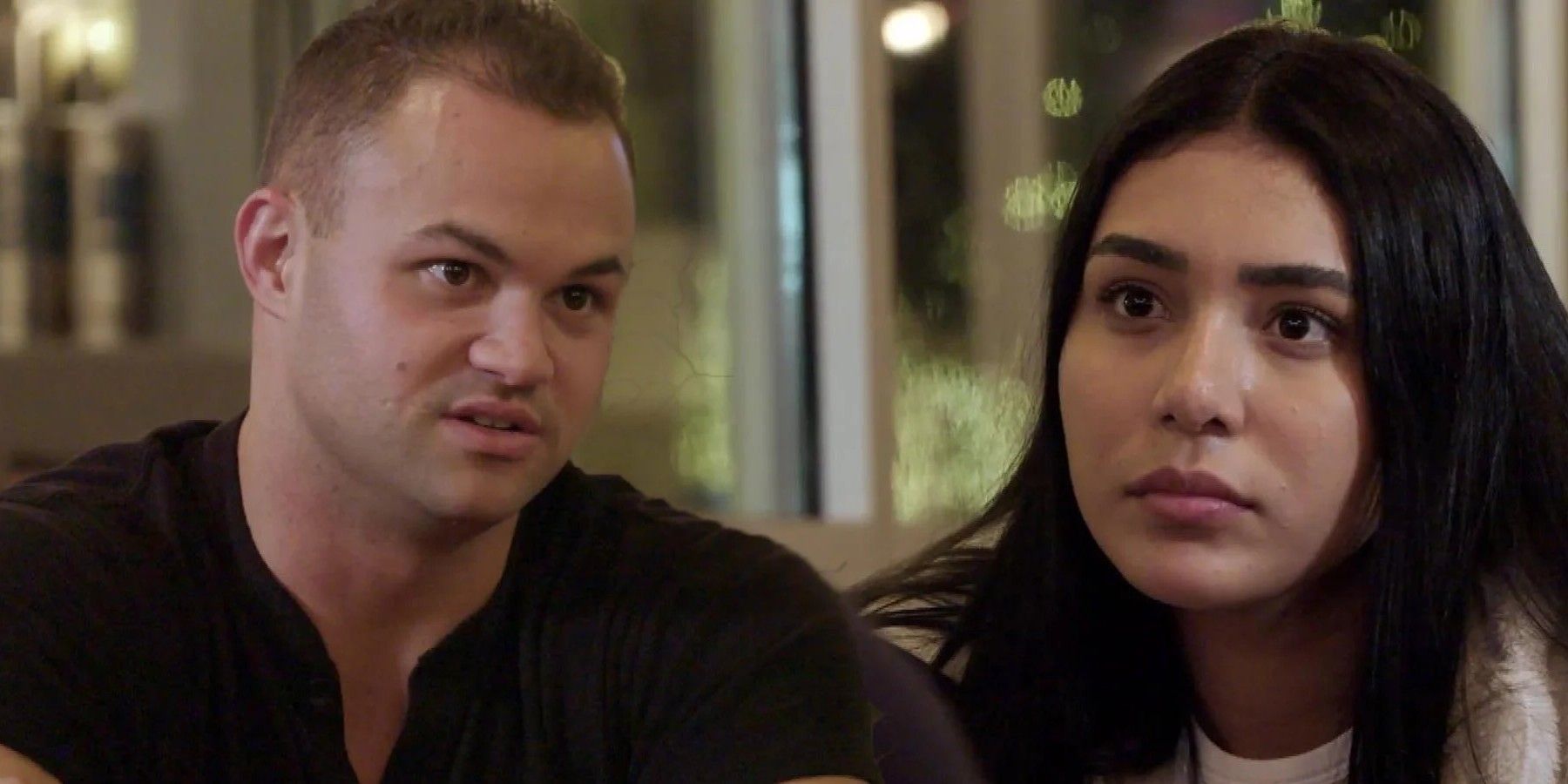 90 Day Fiance Patrick Mendes wearing black shirt and Thaís Ramone wearing white top have intense expressions