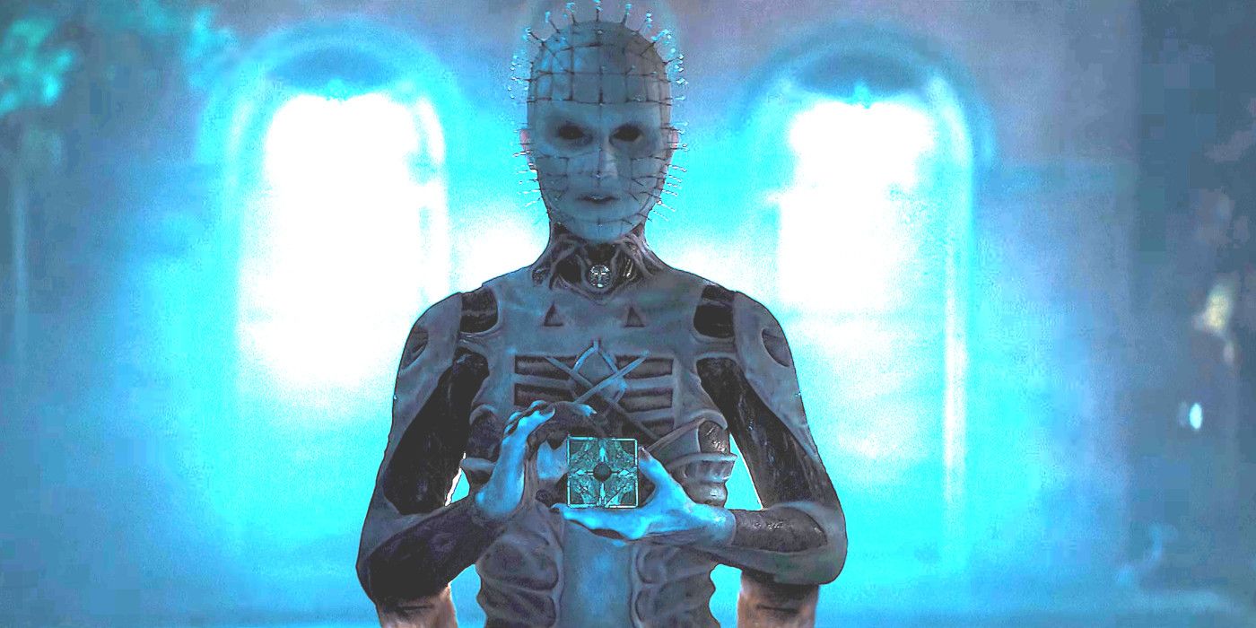 Jamie Clayton como Pinhead em Hellraiser 2022 vestindo uma fantasia horrível com olhos negros segurando um cubo de quebra-cabeça de uma maneira tentadora
