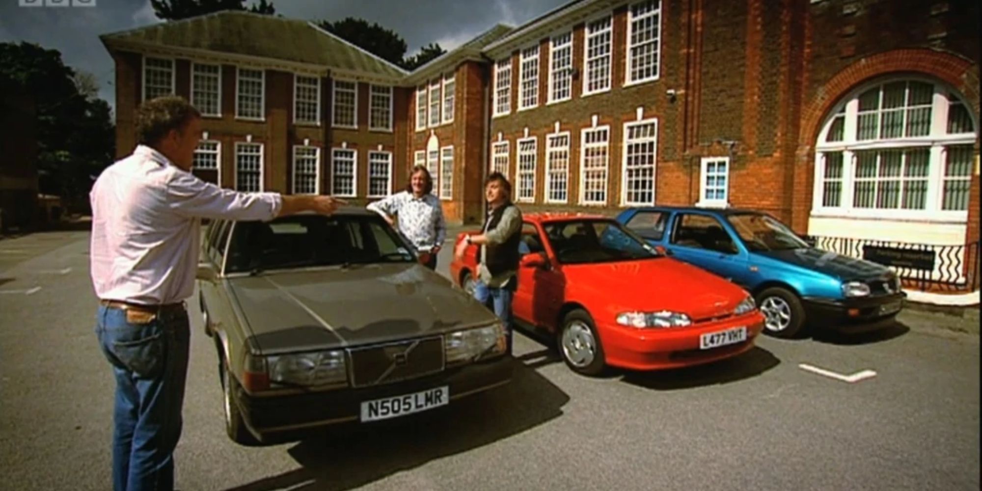 Uma cena de Top-Gear-Season-13, episódio 2 com um carro verde oliva, um carro vermelho e um carro azul em frente a um prédio de tijolos, um membro do elenco apontando
