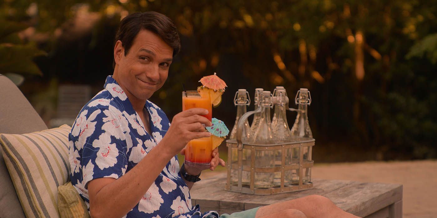 Daniel LaRusso sitting by his pool in a Hawaiian shirt, smiling and holding drinks with umbrellas in a scene from Cobra Kai.