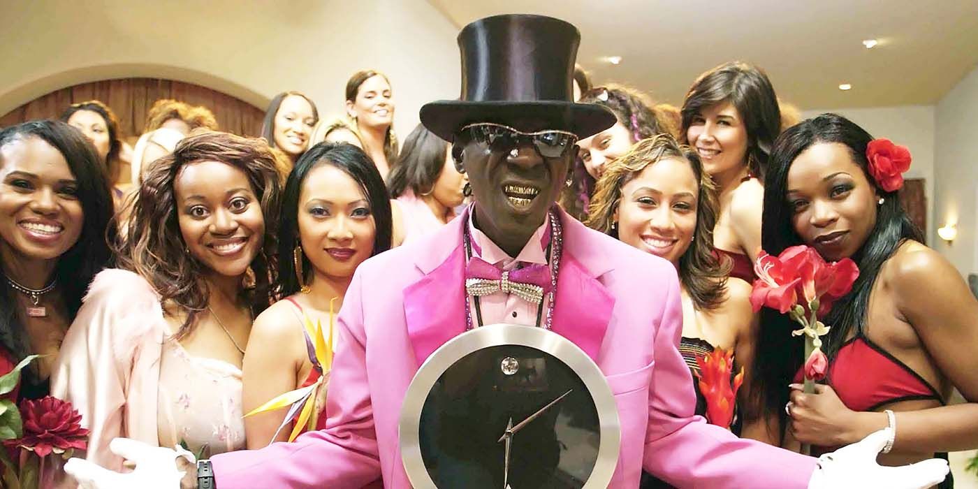 Flavor Flav wearing a top hat in a pink suit standing in front of a group of women in a scene from Flavor of Love.