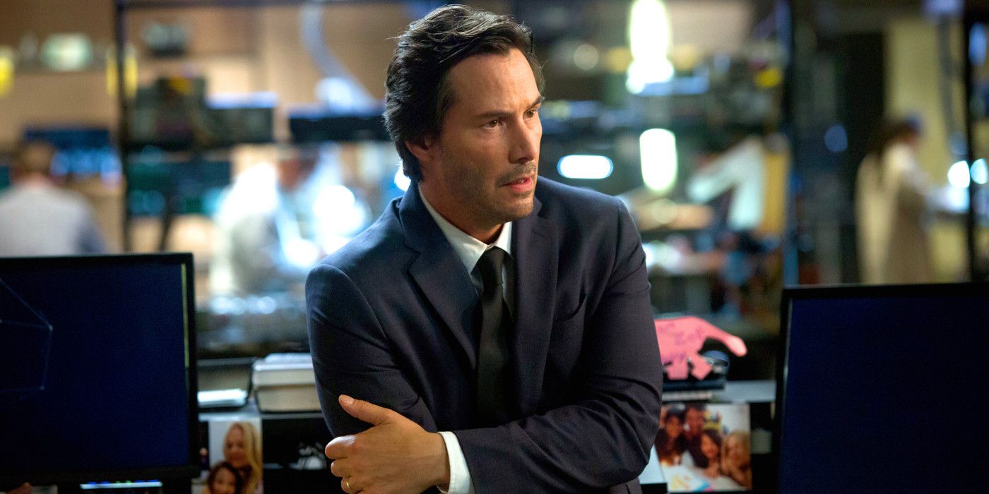 Keanu Reeves in character in the movie Replicas in a business suit before a desk full of computers with his arms folded and a quizzical look on his face