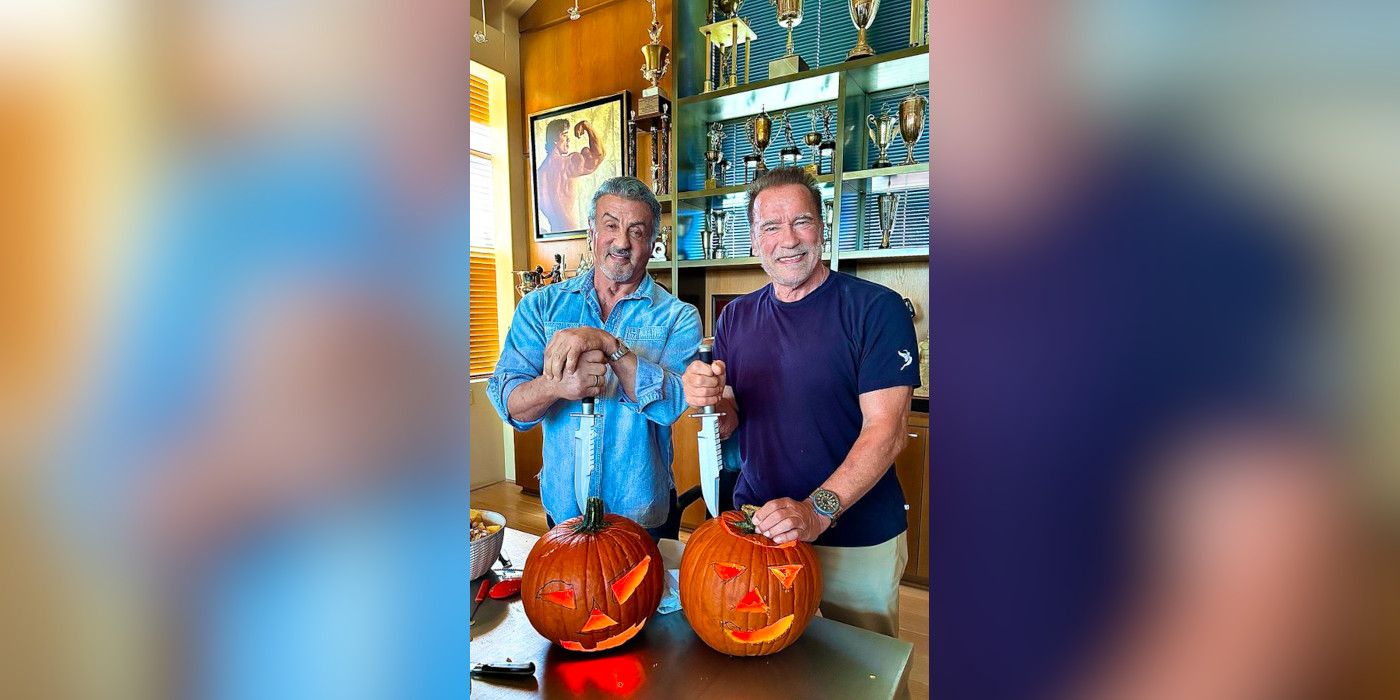 Sylvester Stallone and Arnold Schwarzengger standing together in a trophy room smiling and holding large knives in front of a pair of carved jack-o-lanterns