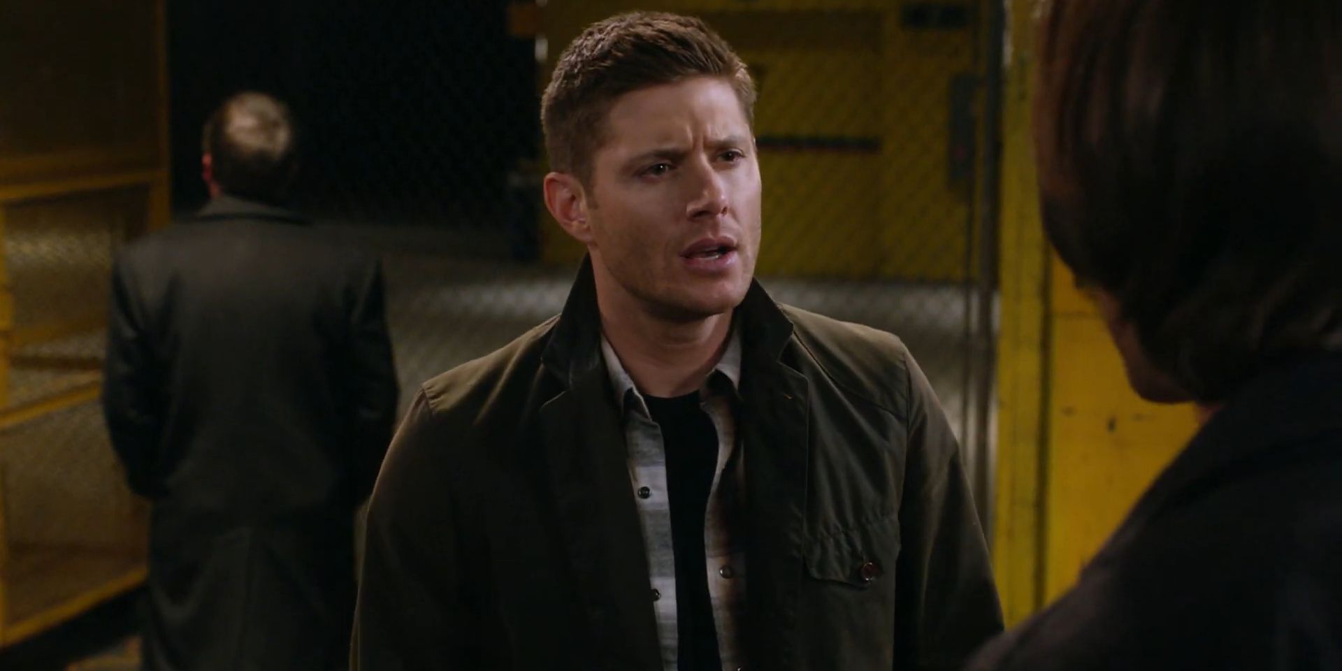 Image of Dean talking to Sam in a warehouse with Crowley in the background
