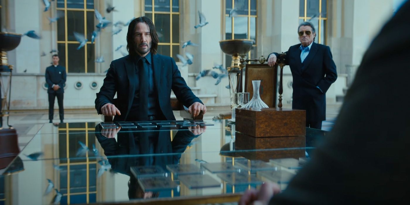 John Wick sitting at a table while Winston stands behind him.
