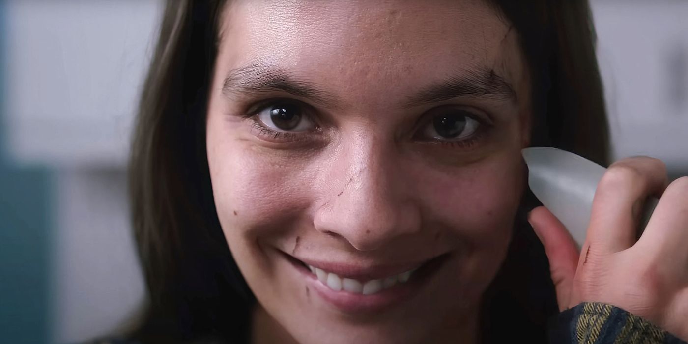 A woman smiling widely while pressing a shard of glass to her cheek in Smile 2022