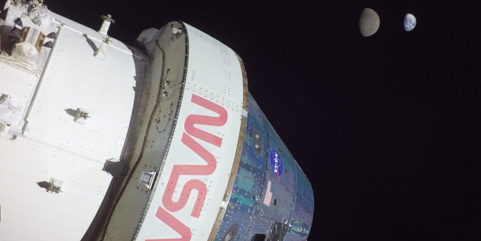 A portion of the Orion spacecraft is pictured in the left side of the image with the NASA logo emblazoned on it in red, while the moon and Earth can be seen at the top right, far in the distance, against the blackness of space