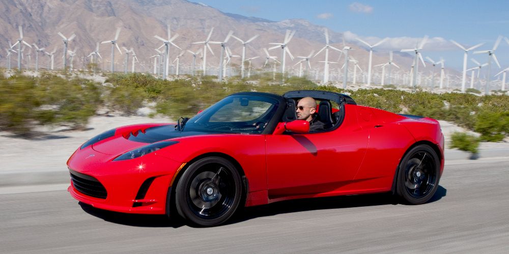 A red Tesla Roadster drives by windmills