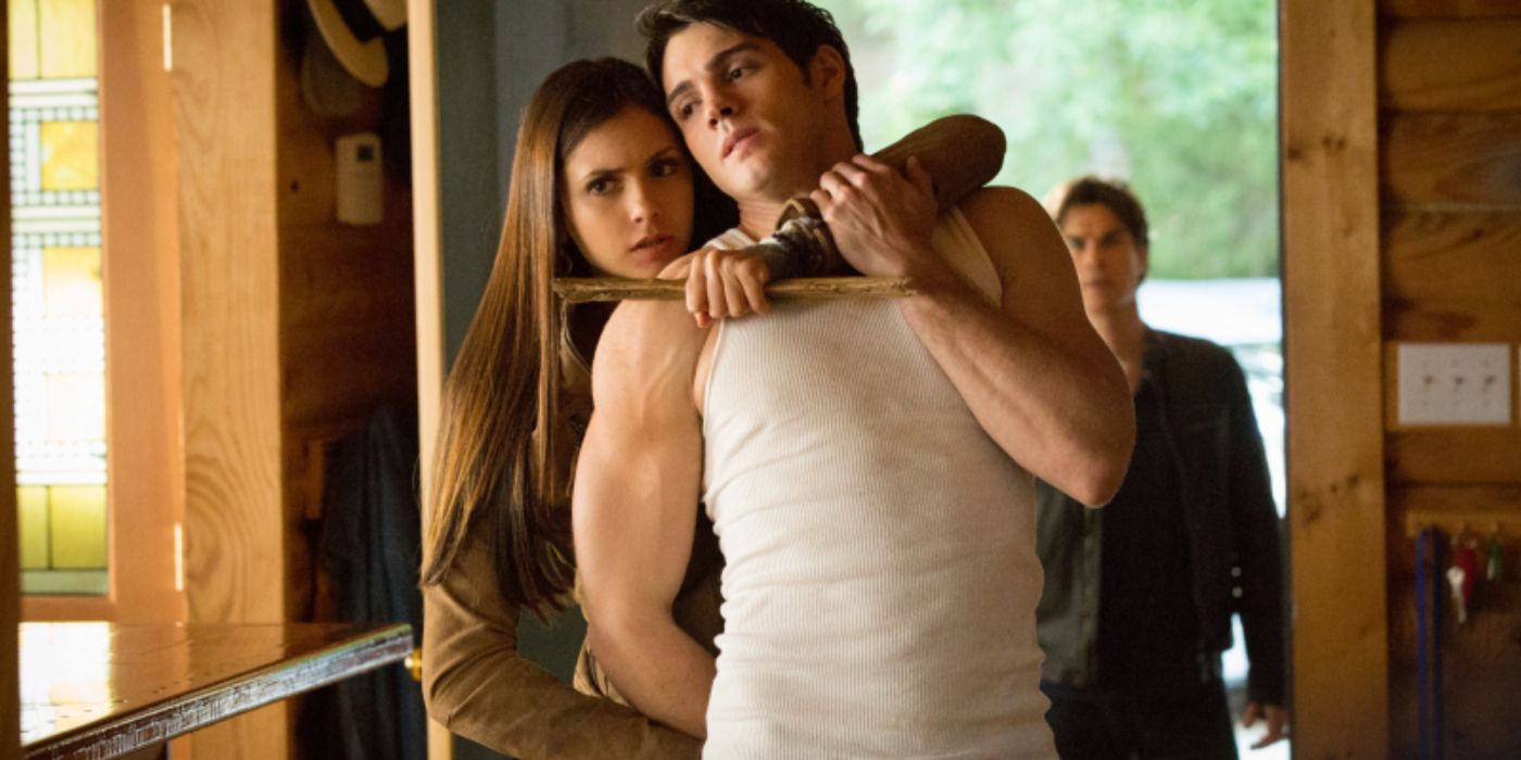 Elena holding her brother Jeremy captive with a wooden stake to his neck with Damon in the background in a Christmas episode of The Vampire Diaries. 