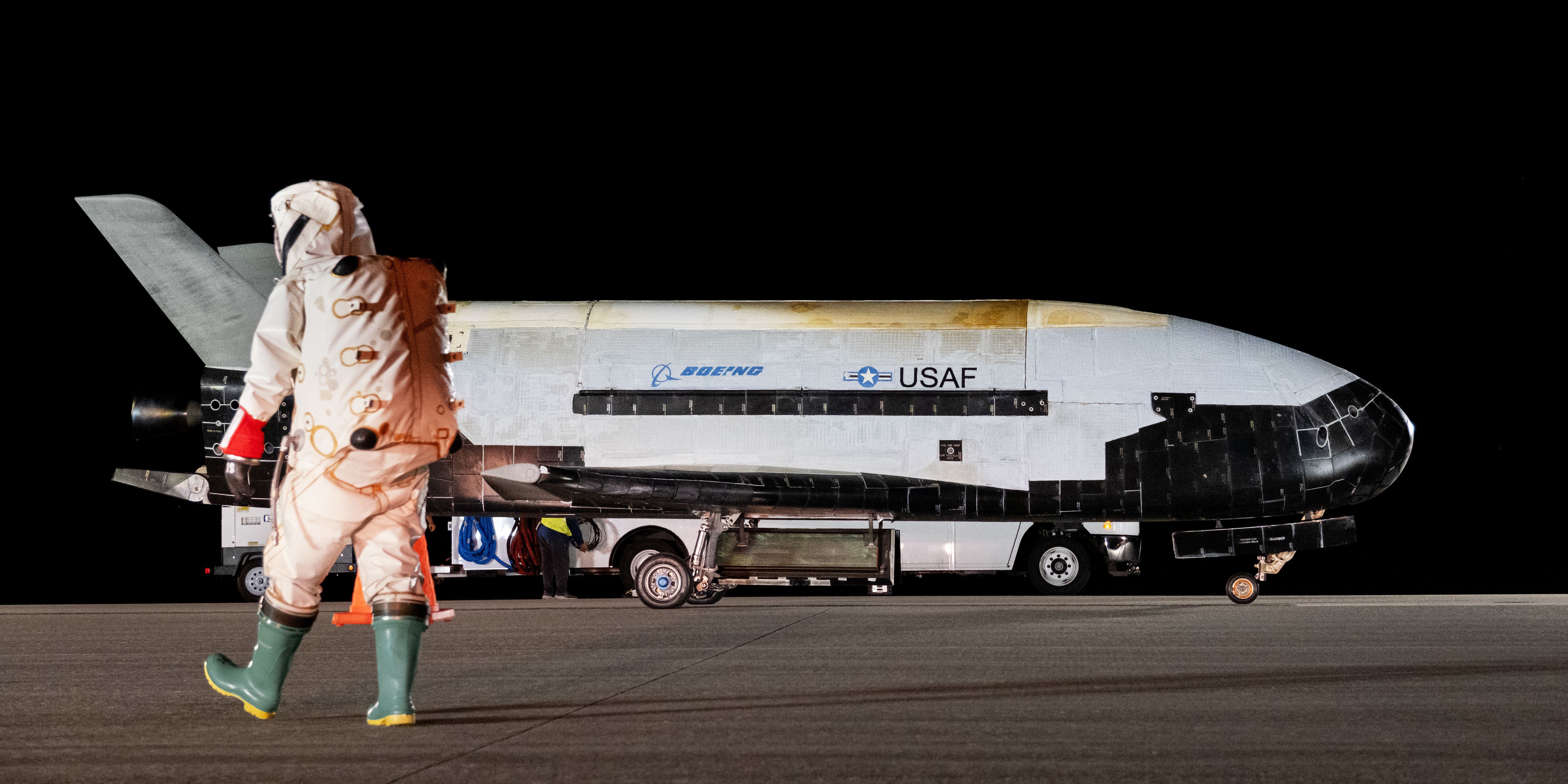 The X-37B spaceplane is pictured on the runway in the background while a person in a white hazmat suit walks past it in the foreground